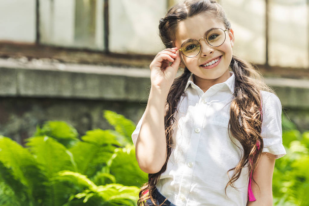 beautiful schoolgirl in uniform and eyeglasses looking at camera in park - Foto, afbeelding