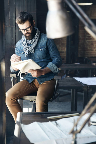 Serious man architect with dark hair and beard, weared in denim jacket, scarf and eyeglasses is sitting in the modern office and designs a new project - Photo, image