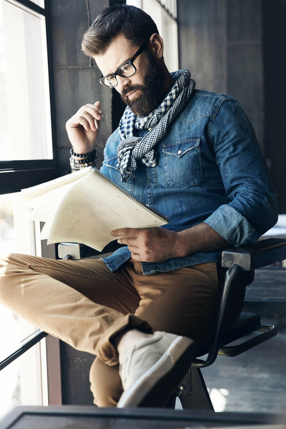 Serious male journalist with dark hair and beard, weared in denim jacket, scarf and eyeglasses is sitting in the modern office and reading his notes  - Φωτογραφία, εικόνα