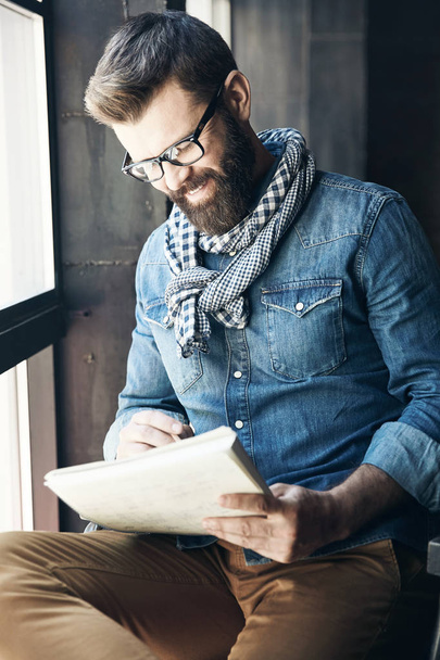 Smiling male writer journalist with dark hair and beard in denim jacket and eyeglasses is sitting in public place and creating new article - Foto, Imagen