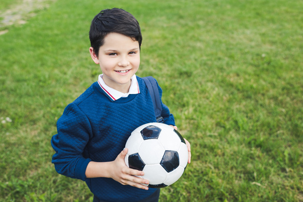 šťastné dítě drží fotbalový míč a při pohledu na fotoaparát na travnaté hřiště - Fotografie, Obrázek