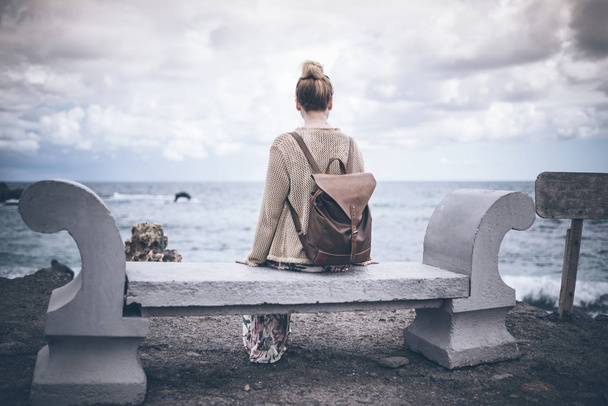 Вид сзади молодой женщины в длинном платье и с рюкзаком, сидящей на каменной скамье у моря
 - Фото, изображение
