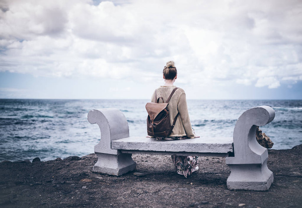Вид сзади молодой женщины в длинном платье и с рюкзаком, сидящей на каменной скамье у моря
 - Фото, изображение