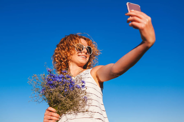 Portrait of beautiful happy woman with red curly hair holding flowers and taking selfie against the blue sky. Having fun during summer holidays - Photo, Image