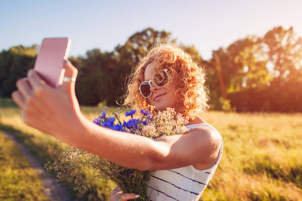 花を押しながら selfie を取って赤巻き毛を持つスタイリッシュな若い女性の屋外のポートレート。夏のレジャー. - 写真・画像