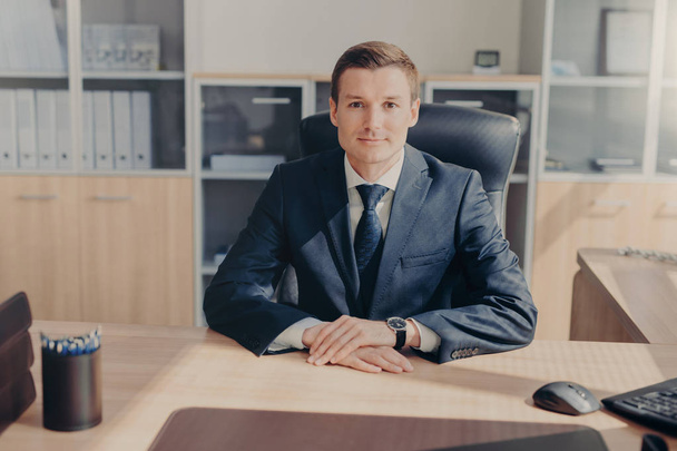 Portrait of attractive prosperous businessman wears black suit, white shirt with tie, sits in his own cabinet, waits for business partners to start meeting, discussing contract terms. Office interior - Foto, Imagem