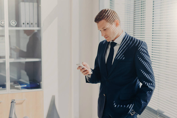 Photo of office employee in elegant outfit, has appealing appearance, uses modern cell phone, checks notification, poses in spacious cabinet, connected to wireless internet. Networking concept - Valokuva, kuva