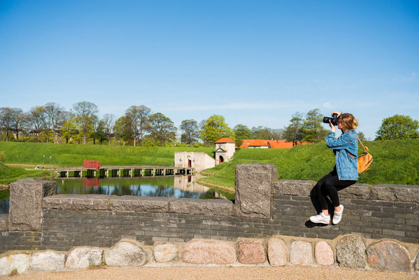 young woman with camera sitting on stone fence and photographing Citadel, copenhagen, denmark - Photo, Image