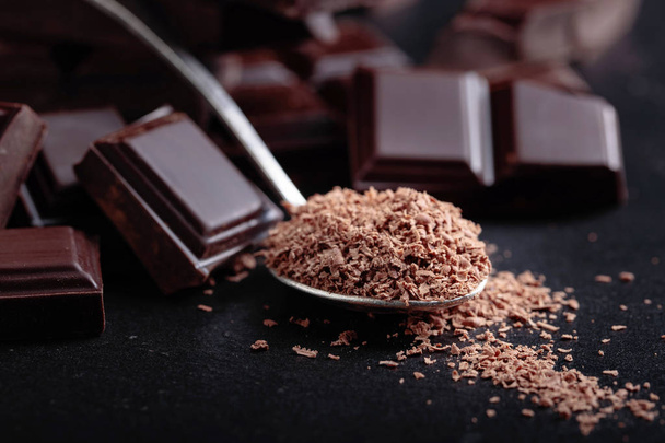 Pezzi di cioccolato nero rotti e gocce di cioccolato in piccolo cucchiaio su sfondo ceramica nera
. - Foto, immagini