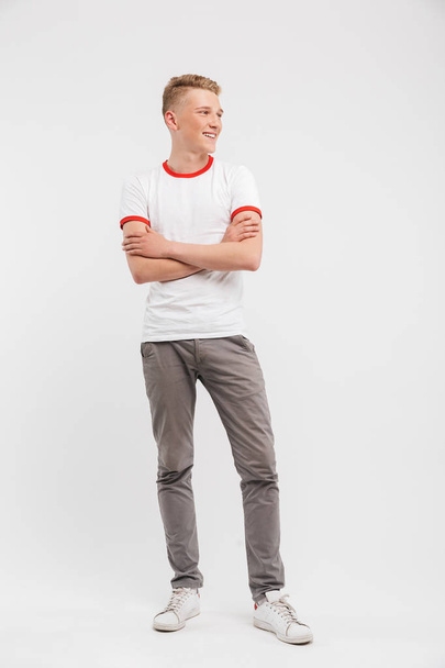 Full length portrait of a smiling teenage boy standing with arms folded and looking away isolated over white background - Zdjęcie, obraz