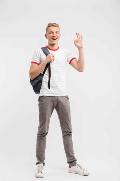 Full length portrait of student boy wearing casual clothing and backpack gesturing alright or ok sign at camera isolated over white background - Foto, Bild