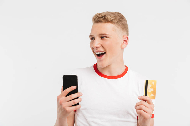 Portrait of an excited teenage boy holding mobile phone and credit card isolated over white background - Фото, изображение