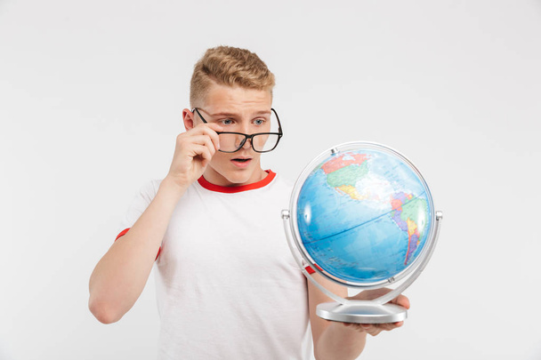 Portrait of a pensive teenage boy in eyeglasses looking at a globe isolated over white background - Foto, Bild