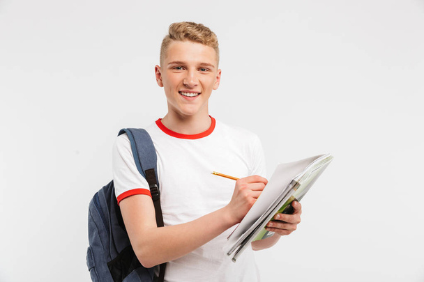 Image of handsome student guy wearing backpack smiling and looking at you while writing down notes at textbook isolated over white background - Фото, зображення