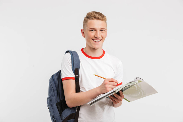 Image of attractive university or college student wearing backpack smiling and looking at you while writing down notes at textbook isolated over white background - Foto, Bild