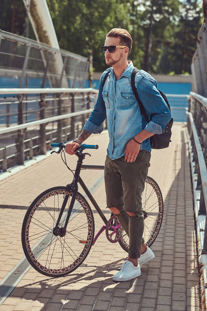 Handsome fashionable hipster with a stylish haircut in sunglasses walking with bicycle outdoors. - Fotó, kép