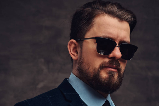 Close-up portrait of a handsome fashionable middle-aged man with beard and hairstyle dressed in an elegant formal suit and sunglasses on a textured dark background in studio. - Photo, Image