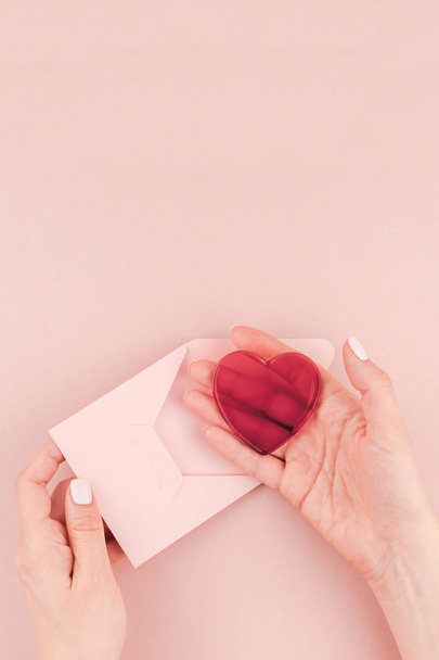 Creative image of woman hand holding small love letter  and red heart with copy space on millennial pink background in minimalism style. Concept template for feminine blog, social media - Foto, Bild