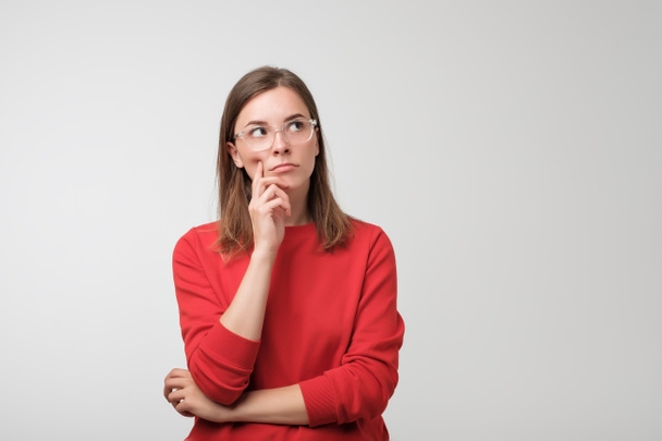 pensativa mujer europea en suéter rojo está mirando hacia arriba pensando o planeando
. - Foto, Imagen