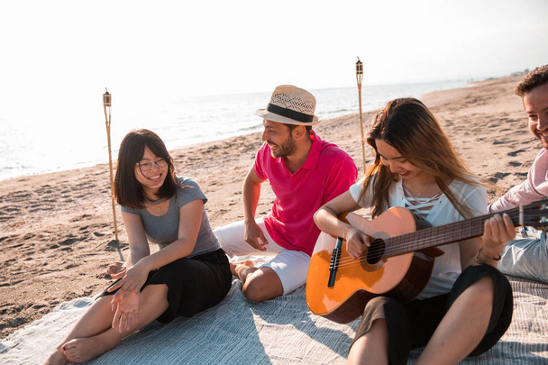 Happy multiracial young people playing guitar and making picnic at the beach. Friends, music and holiday concept. - Foto, Imagem