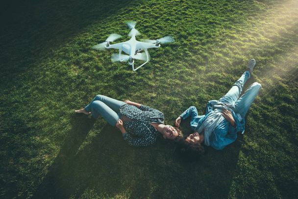 zwei wunderschöne, lockige Mädchen verschiedener Rassen liegen auf dem vom Sonnenlicht erhellten Rasen der Sommerlichtung und zeichnen mit einer fliegenden Drohne über ihnen eine Sendung für ihre Vlogs auf - Foto, Bild