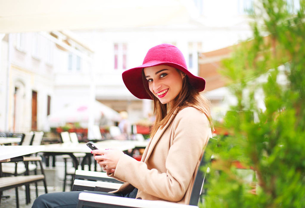 Lachende meisje in een jas zitten in een straat Cafe in het stadsplein met een smartphone - Foto, afbeelding