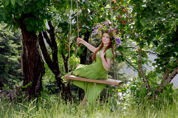 Young, beautiful woman in a green dress with a wreath of wild flowers sits on a homemade swing in a mountain garde - Photo, Image