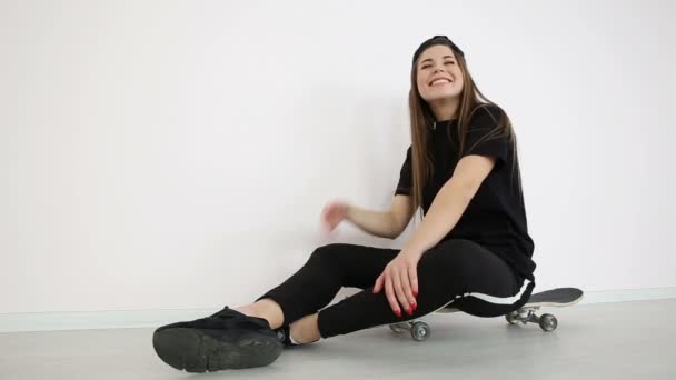 dospívající dívka v módní hip hop oblečení a čepici vystupují proti bílé zdi se skateboard - Záběry, video