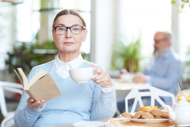 Elegante vrouw in brillen en smart casual leesboek terwijl het hebben van thee met cookies in café - Foto, afbeelding