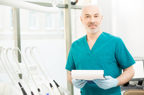 Alter Zahnarzt in Uniform und Handschuhen blickt in die Kamera, während er mit Zahnarztgeräten an seinem Arbeitsplatz steht - Foto, Bild