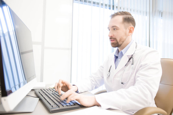 Jeune homme en whitecoat assis par moniteur d'ordinateur à son bureau dans les cliniques et de consultation des patients en ligne
 - Photo, image