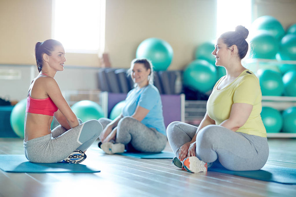 Twee vrouwen en hun fitness instructeur overgewicht met gekruiste benen zit op matten en bespreken van oefeningen voor training - Foto, afbeelding