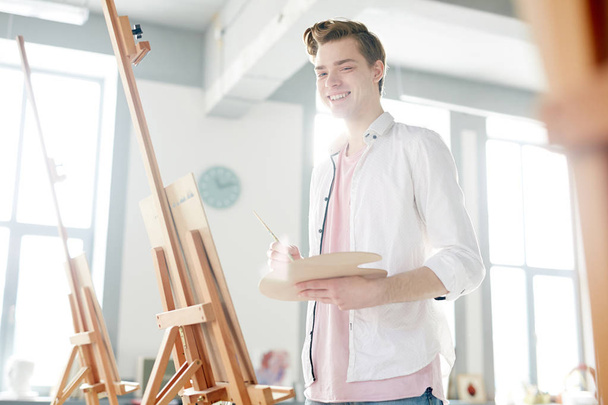 Happy guy with palette looking at camera while standing by easel in classroom - Foto, Imagem