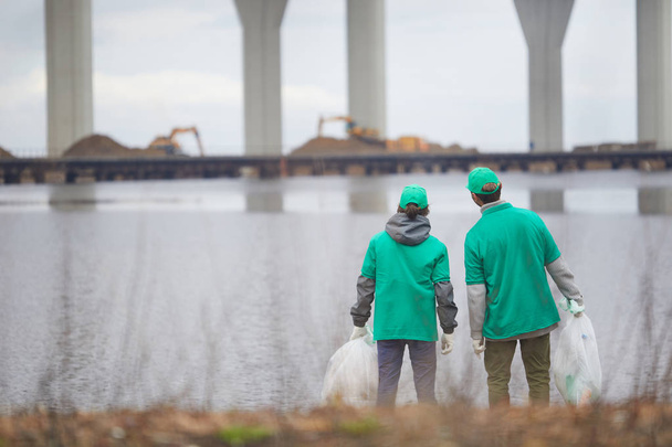 Οπίσθια όψη της greenpeace άνδρες με σάκοι απορριμάτων στέκεται από waterside σε αστικό περιβάλλον - Φωτογραφία, εικόνα