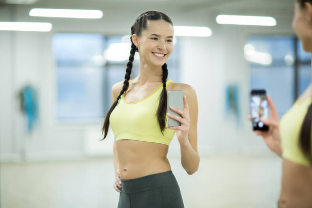 Fit young woman in activewear looking at herself in front of mirror and making selfie in sports center - Photo, image