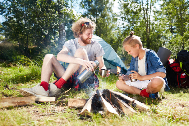 Young man and cute boy with hot drinks sitting by bonfire after breakfast in the morning during their trip - Photo, Image