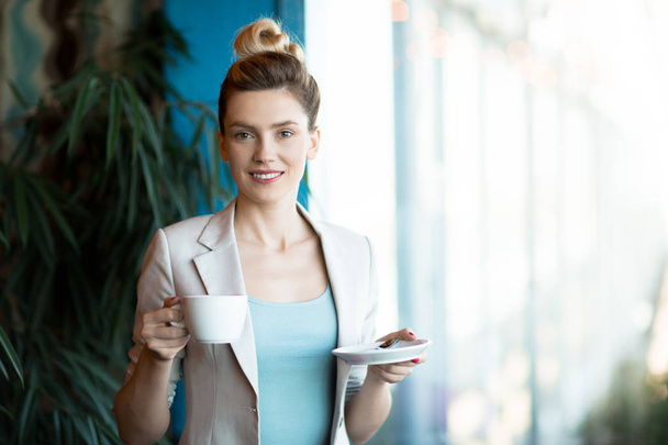 Young female manager with cup of tea or coffee and saucer looking at camera during tea-break in office - Foto, afbeelding
