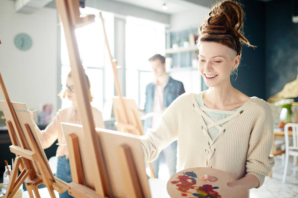 Radostné teenager s barvami palety při pohledu na co malovala na stojanu během lekce - Fotografie, Obrázek
