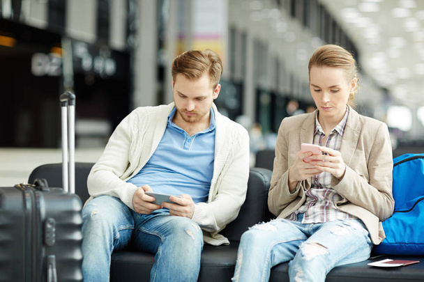 Два современных мужчины и женщины в повседневной одежде сидят в зале аэропорта и ищут в своих смартфонах
 - Фото, изображение