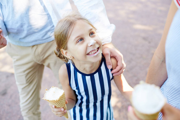 lustiges Mädchen mit Eis lächelt ihre Mutter an, während sie an einem sonnigen Tag mit ihren Eltern spazieren geht - Foto, Bild