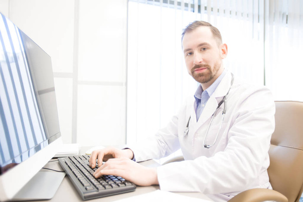 Молодой врач смотрит в камеру, сидя за монитором компьютера во время онлайн-консультации
 - Фото, изображение