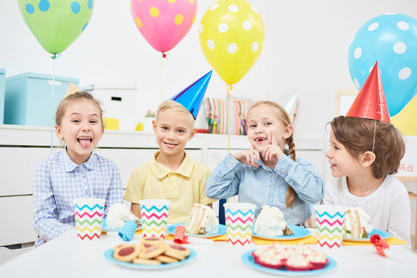 Niños divertidos en gorras de cumpleaños disfrutando de la fiesta con globos y sabrosos postres en el jardín de infantes
 - Foto, imagen
