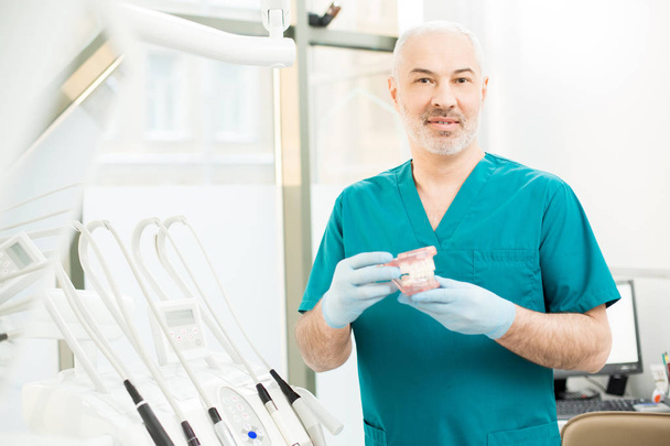 Dentista profissional em uniforme mostrando dentes falsos enquanto trabalhava em clínicas odontológicas modernas
 - Foto, Imagem