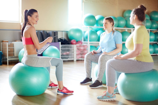 Молодий інструктор з фітнесу та її група сидять на зелених фігурних м'ячах у спортзалі та обговорюють, які вправи вони збираються робити
 - Фото, зображення