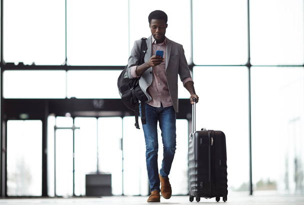 Jonge mobiele reiziger met smartphone, rugzak en koffer wandelen langs luchthaven en messaging of kijken door on line informatie - Foto, afbeelding