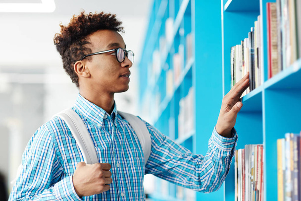 Jonge college student permanent door boekenplank en op zoek naar degene die is te lezen voor de volgende les - Foto, afbeelding