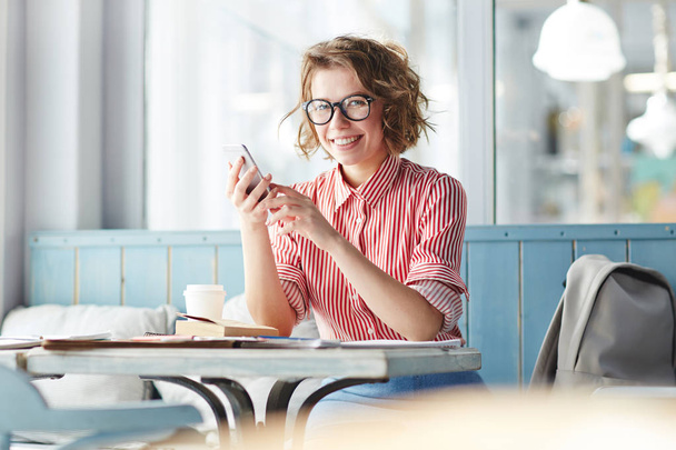Chica feliz mirando a la cámara mientras se sienta junto a la mesa en el café y compartir sus fotos con amigos en las redes sociales
 - Foto, Imagen