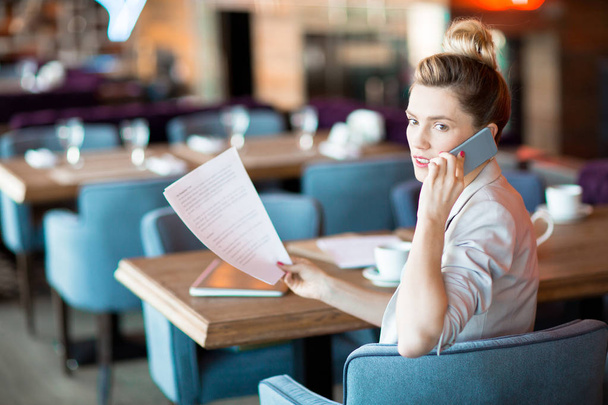 Jeune femme occupée avec smartphone et papiers financiers assis dans un fauteuil dans un café ou un restaurant moderne
 - Photo, image