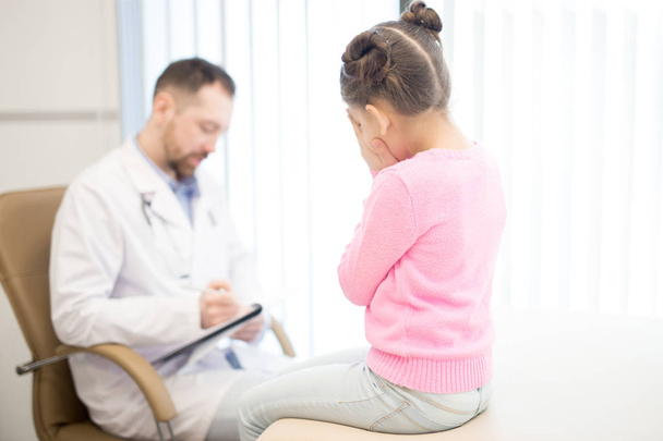 Little girl is afraid of bad diagnosis while pediatrician making prescriptions in her medical card - Fotografie, Obrázek