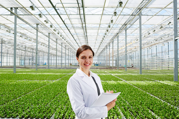 Jonge succesvolle bioloog in whitecoats op achtergrond van grote groene plantage in kassen - Foto, afbeelding